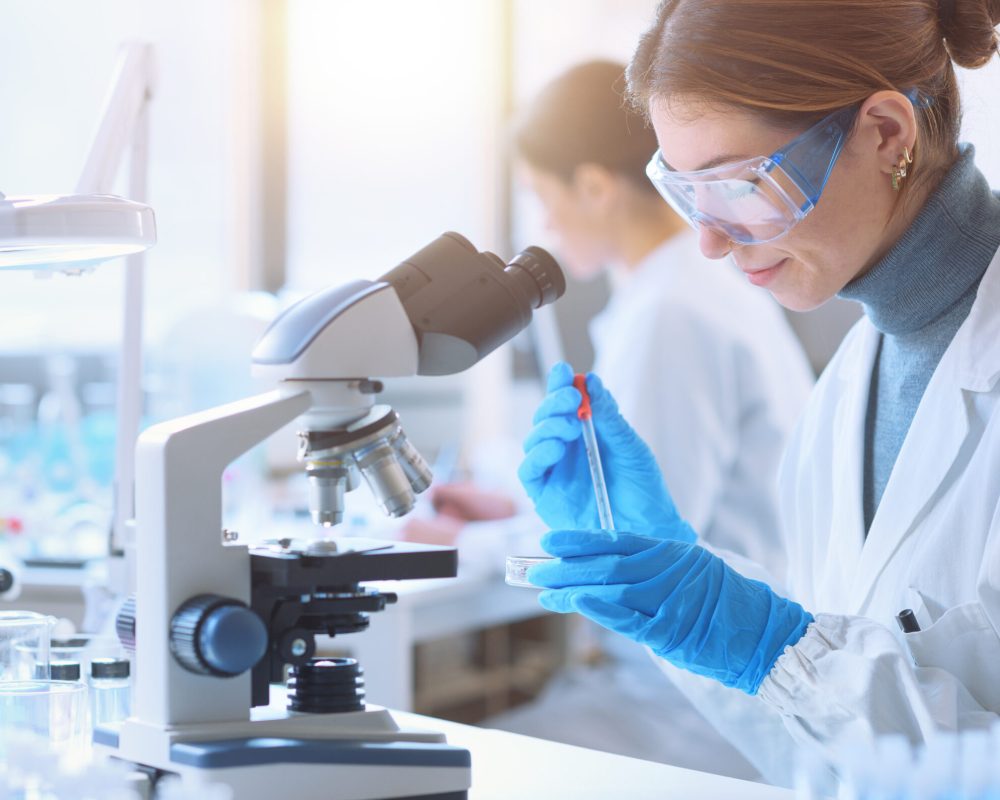 Young female student in the research lab, she is examining a sample in a petri dish and using the microscope