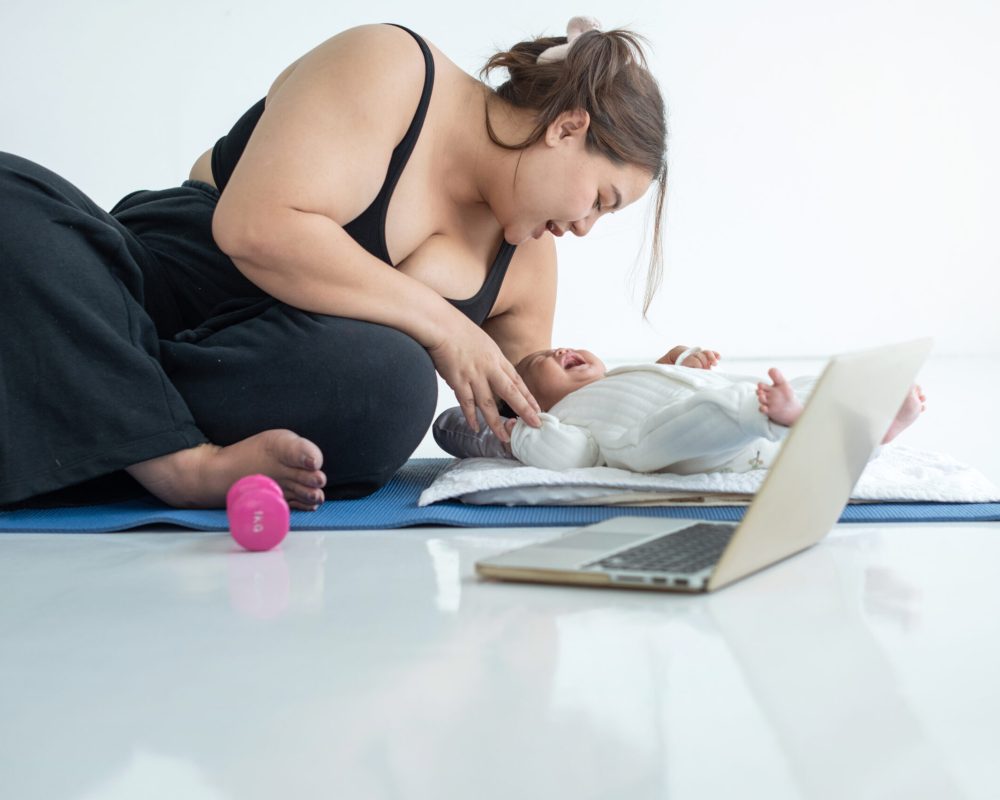 Chubby mom in sportswear pause  her online fitness class on laptop and comfort her crying baby, doing exercise on floor mat at home